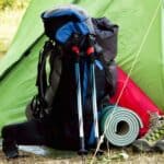 A camping backpack, trekking poles, rolled-up sleeping pad, and hiking boots are positioned in front of a green and red tent set up outdoors.