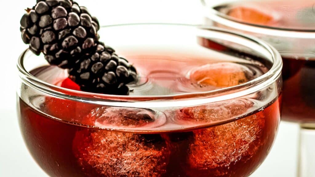 Close-up of a clear glass of red beverage with ice cubes, garnished with a blackberry on the rim. Another similar glass is partially visible in the background.