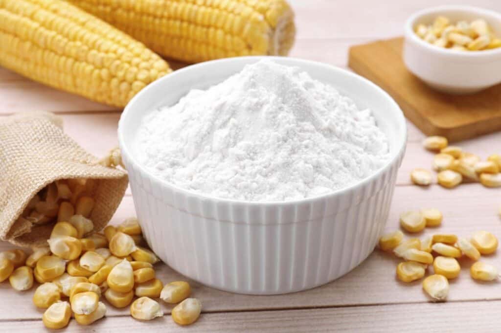 A bowl filled with corn flour, surrounded by corn kernels and two ears of corn in the background.