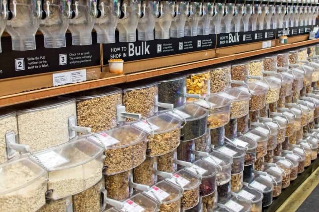 A bulk food section with various dry goods in clear containers, each labeled with product names and codes.