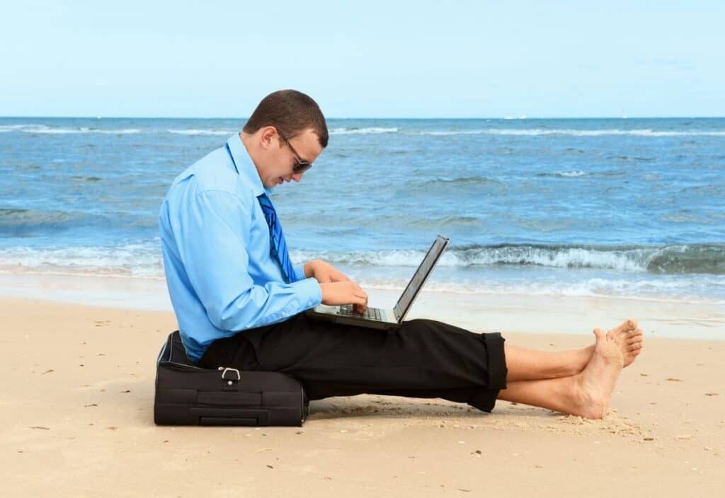 A man in a blue shirt and black pants sits barefoot on a beach, using a laptop placed on his lap. He is seated on a black briefcase, with the ocean in the background.