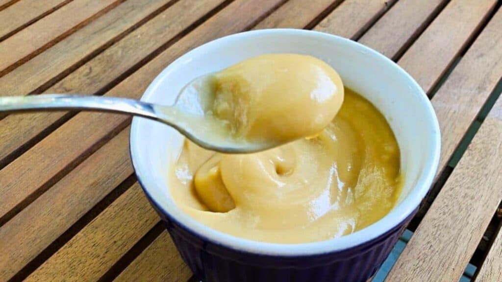 Image shows A spoon lifting a scoop of creamy butterscotch pudding from a small, dark-colored bowl on a wooden table.