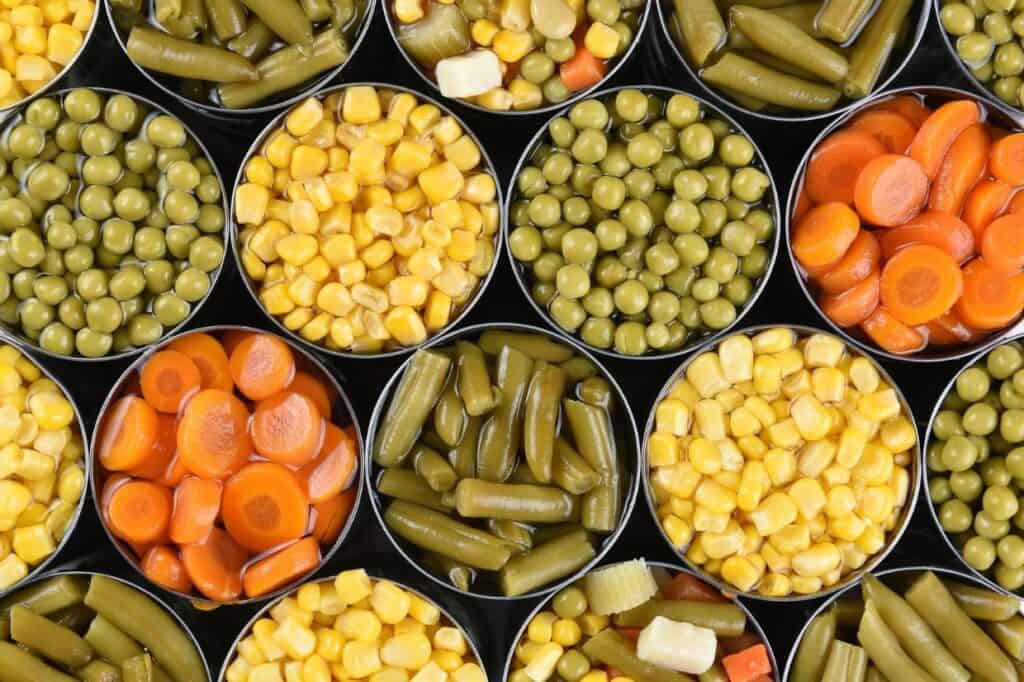 Close-up view of open cans filled with various vegetables, including corn, peas, carrot slices, green beans, and mixed vegetables, arranged in a grid pattern.