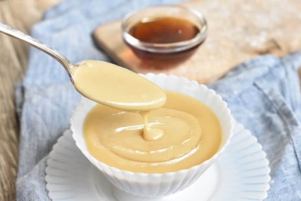 A spoon dipped into a bowl of thick, creamy custard with a small bowl of syrup in the background.
