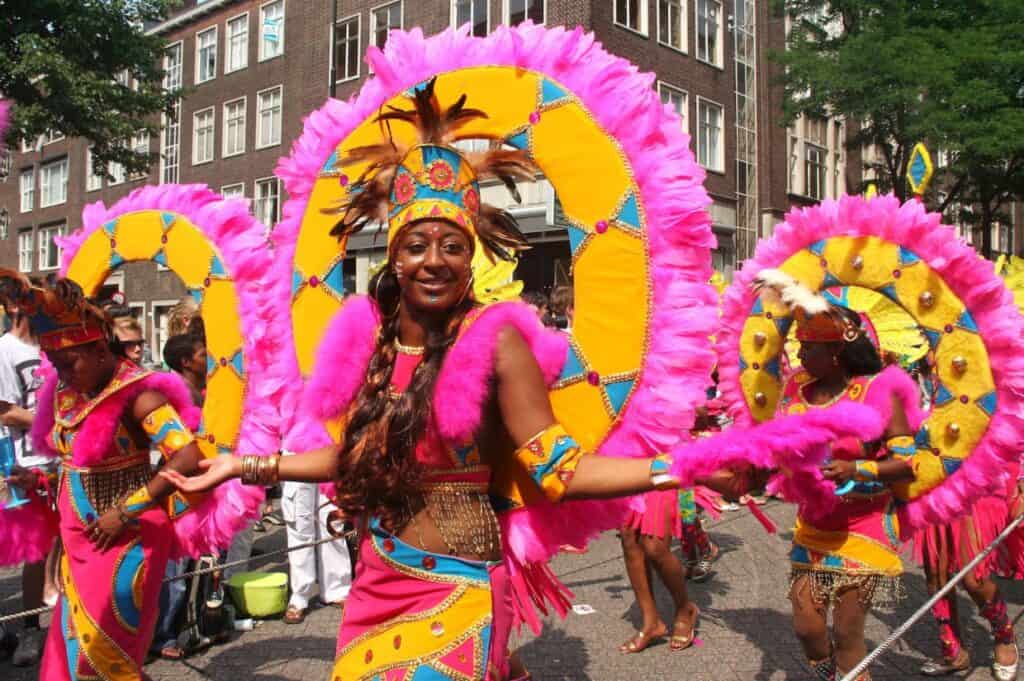 Performers in vibrant, colorful costumes with large feathered headdresses, embodying the essence of Caribbean culture, participate in a street parade. They smile and pose amidst buildings and onlookers.