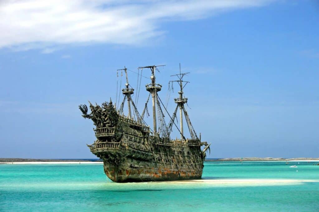 An old, abandoned pirate ship with weathered sails is docked in clear, shallow turquoise waters under a partly cloudy sky, reminiscent of a bygone era of pirates and privateers.