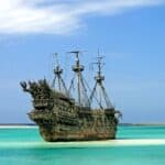 An old, abandoned pirate ship with weathered sails is docked in clear, shallow turquoise waters under a partly cloudy sky, reminiscent of a bygone era of pirates and privateers.
