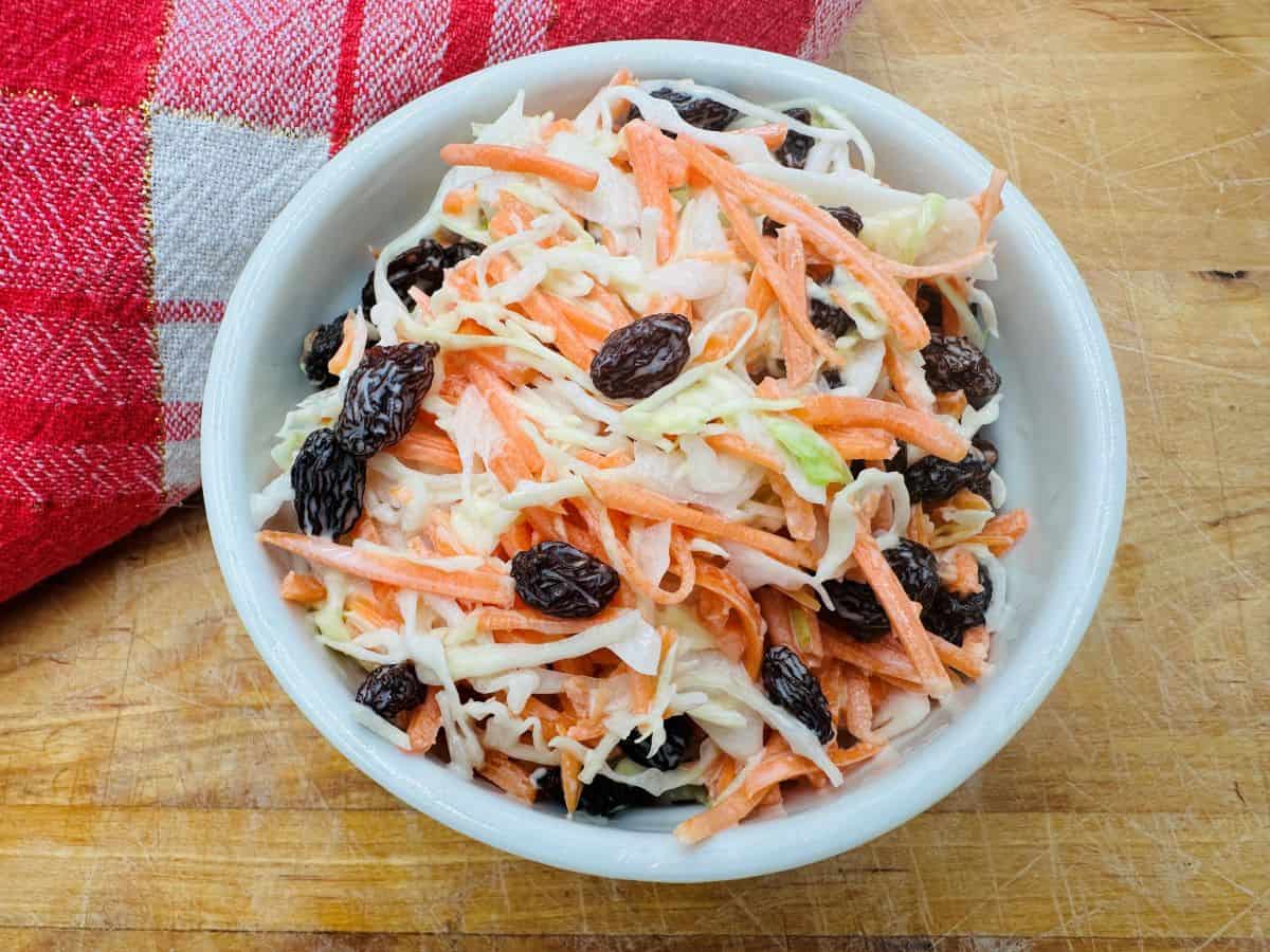 A bowl of coleslaw with shredded carrots and raisins on a wooden surface, next to a red and white checkered cloth.