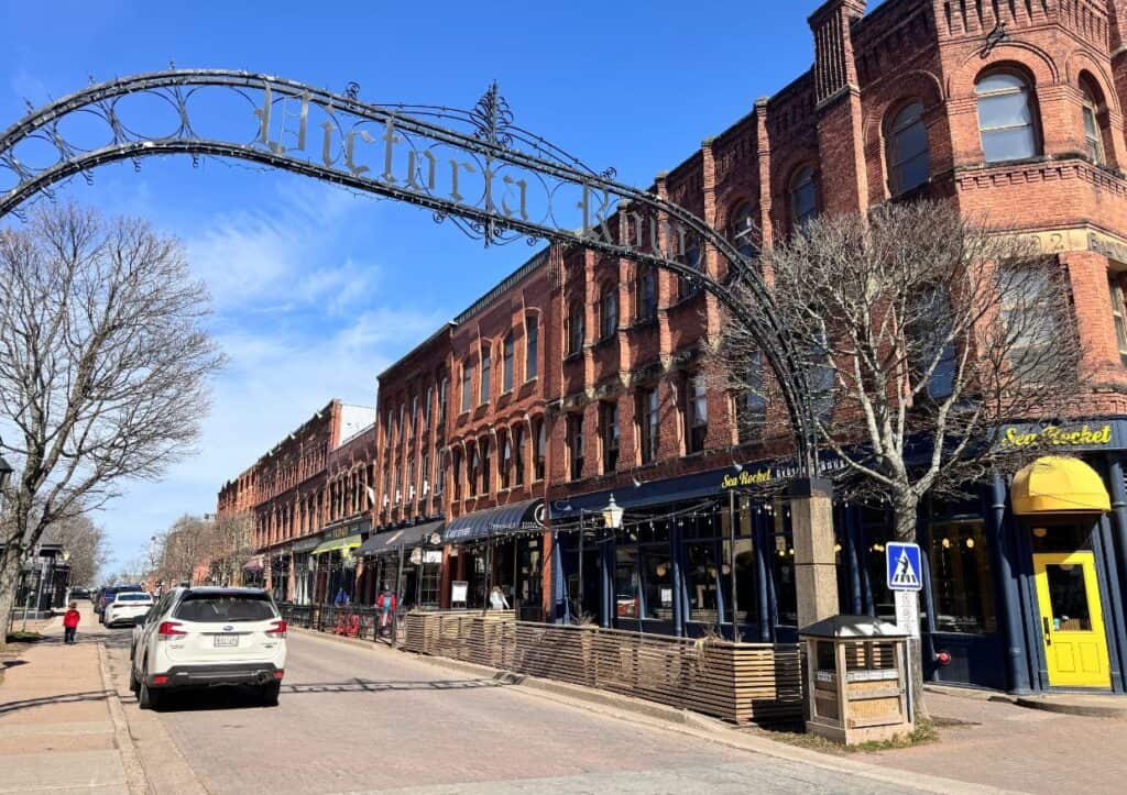 A street scene with historic brick buildings, shops, a wrought iron archway, parked cars, and pedestrians. The weather is sunny with a clear blue sky.