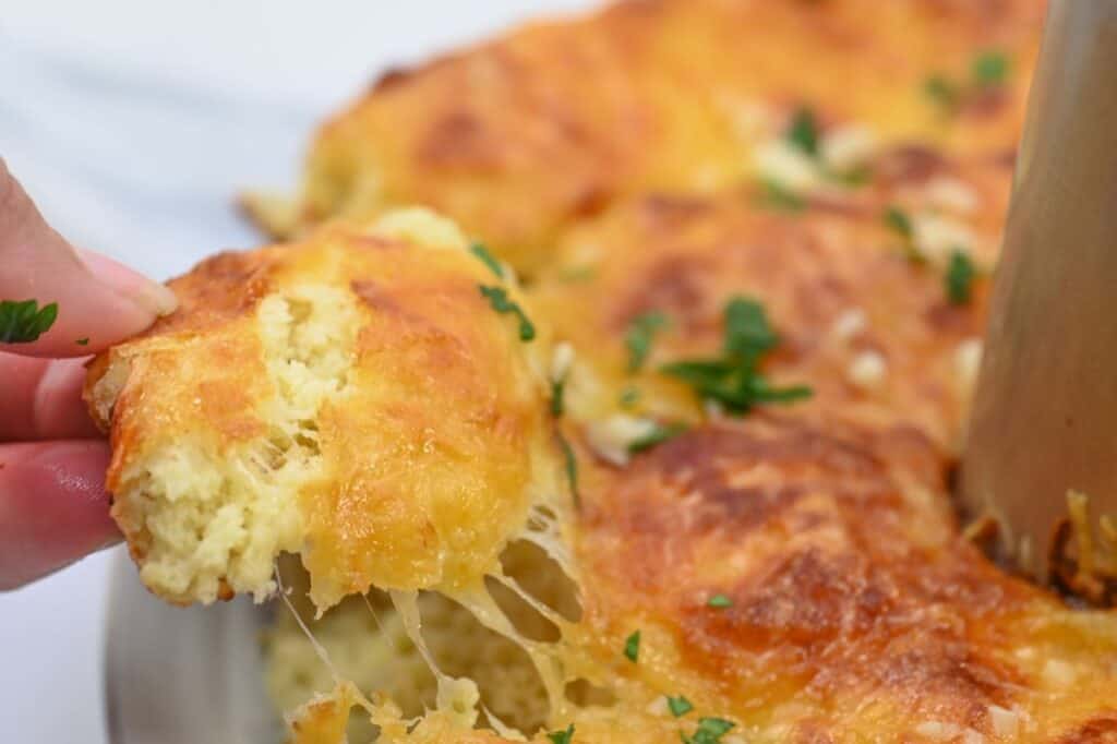 A hand pulls a piece of cheesy bread topped with melted cheese and garnished with chopped herbs from a baking dish.