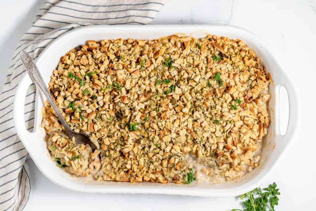 A white casserole dish filled with baked stuffing topped with crushed bread crumbs and garnished with parsley. A serving spoon is placed in the dish. A striped kitchen towel is partially visible.