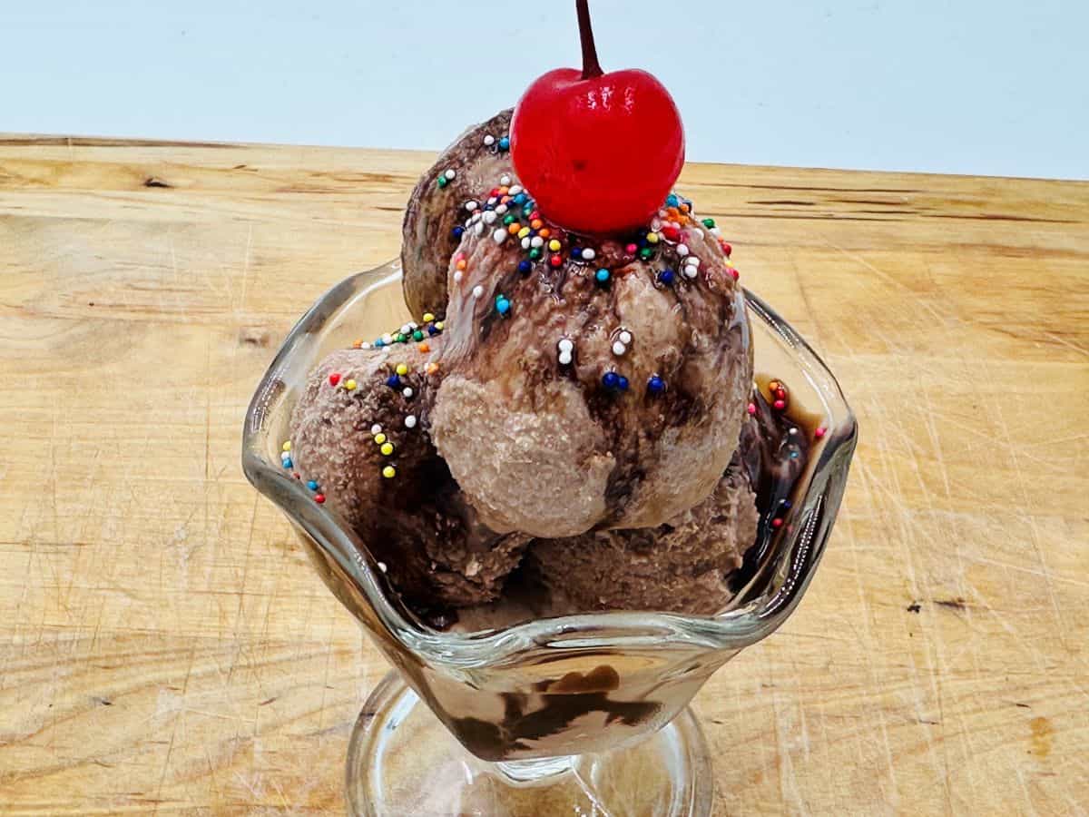 A glass bowl filled with scoops of chocolate ice cream topped with colorful sprinkles and a red cherry, placed on a wooden surface.