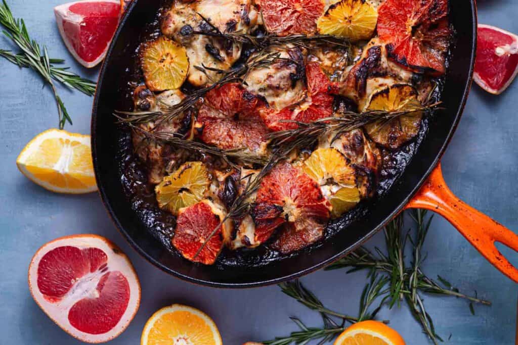 A cast-iron skillet with cooked chicken pieces topped with grilled citrus slices and sprigs of rosemary. Halved and quartered citrus fruits are arranged around the skillet on a blue surface.