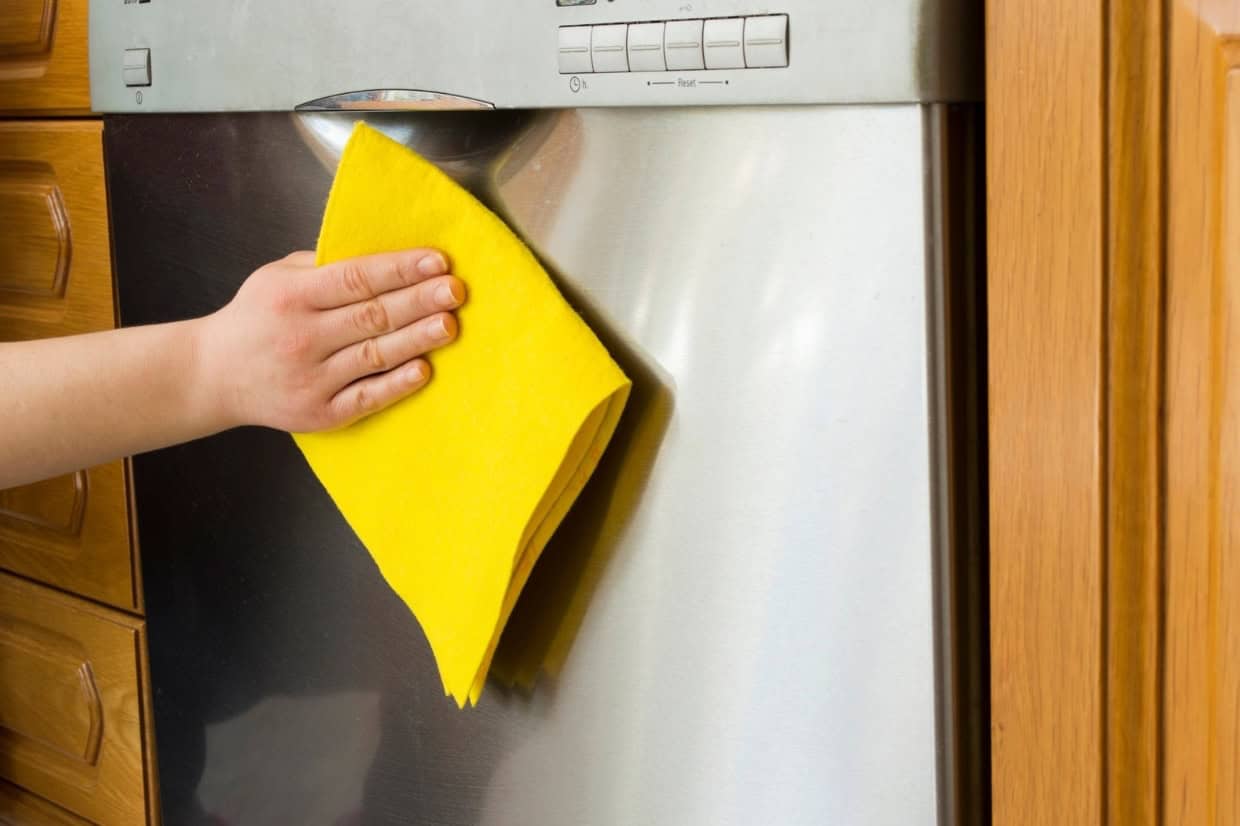 A person is cleaning the stainless steel surface of a dishwasher with a yellow cloth.