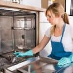 Person wearing gloves and an apron cleans the interior of a dishwasher.