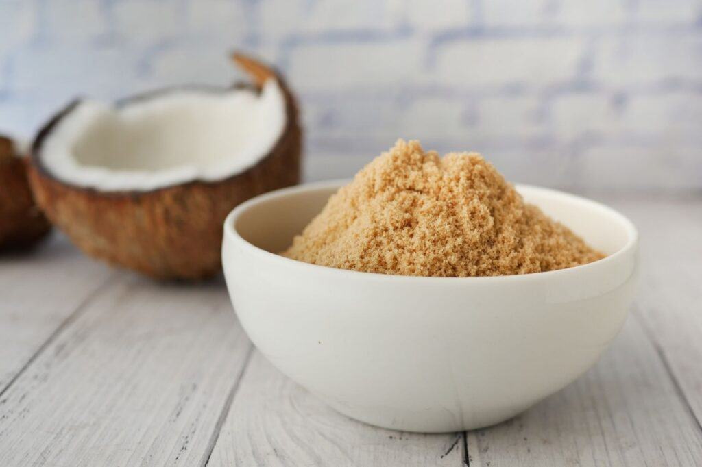 A white bowl filled with coconut sugar is placed on a white wooden surface with a halved coconut in the background.