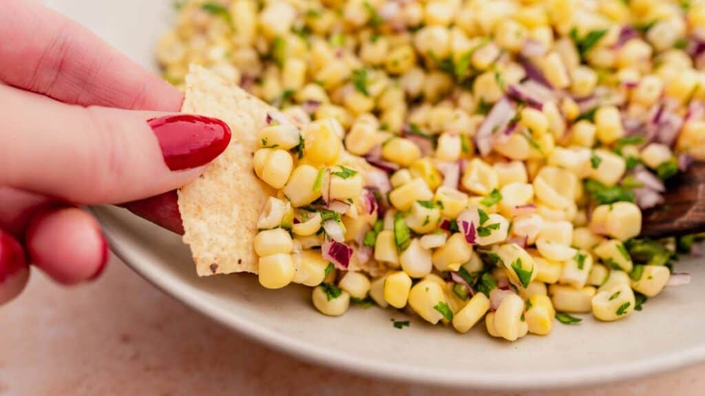 A hand with red nail polish dips a tortilla chip into a bowl of corn salsa, featuring corn kernels, chopped red onion, and cilantro.