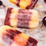 Three multi-colored popsicles, featuring bands of yellow, orange, and purple hues, rest on a bed of ice cubes surrounded by fresh cherries.