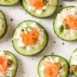 Slices of cucumber topped with a dollop of cream cheese, smoked salmon, capers, and dill, arranged on a white speckled surface.