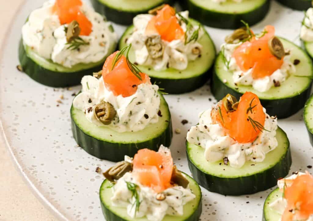 Cucumber slices topped with creamy cheese, smoked salmon, capers, and dill are arranged on a plate.