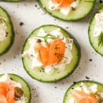 Cucumber slices topped with cream cheese, smoked salmon, capers, and dill, arranged on a white speckled surface.