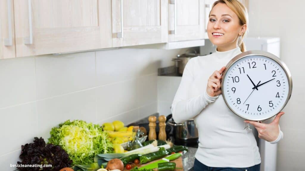 A person in a kitchen holds a large wall clock showing 9:00. A variety of fresh vegetables and fruits are on the counter.