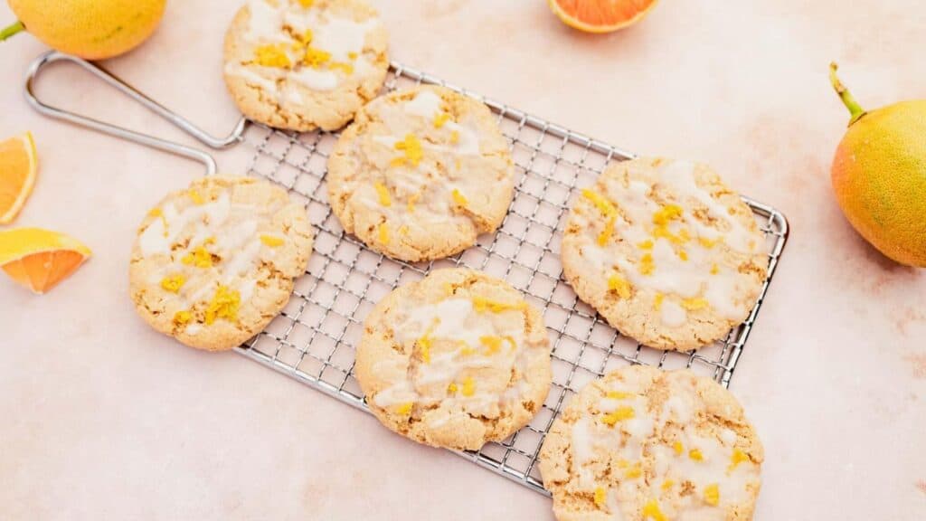 Six lemon zest glazed cookies on a cooling rack with sliced and whole lemon around them on a light surface.