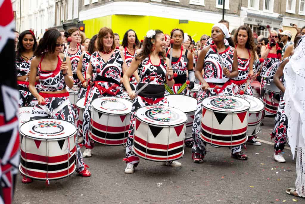 A group of people in matching red, white, and black attire play large drums on a city street, their vibrant rhythms bringing the vibrant essence of Soca to Calypso, surrounded by enthusiastic onlookers.