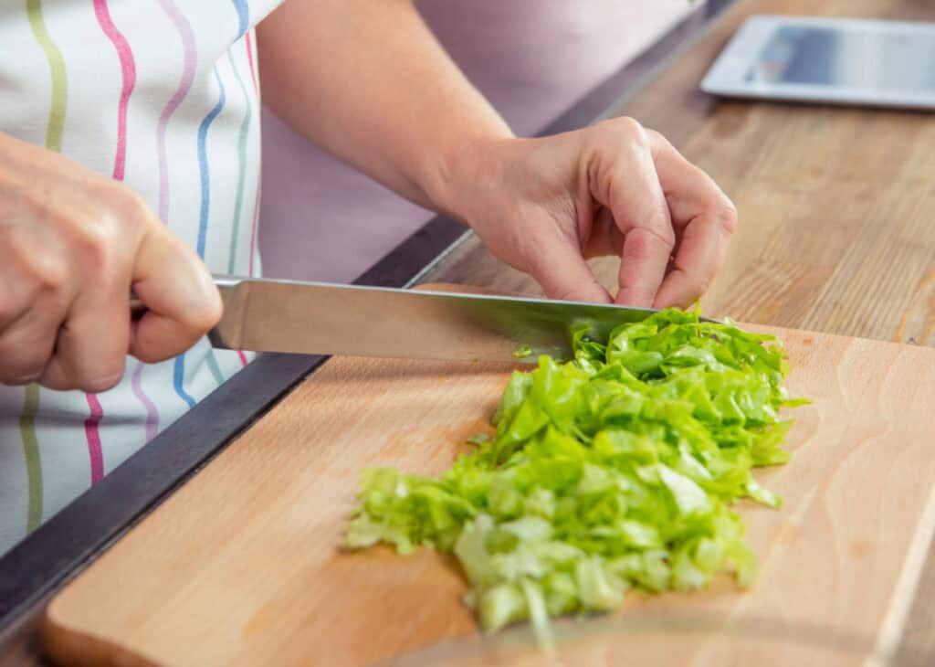 Person chopping green lettuce on a wooden cutting board with a large knife. An electronic tablet is visible in the background.
