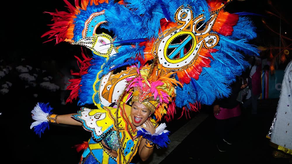 A person dressed in a vibrant and colorful feathered costume smiles and dances during a lively nighttime parade, celebrating Caribbean history and heritage.