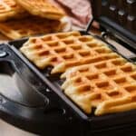 Two freshly cooked waffles inside a waffle maker with more waffles on a plate in the background.