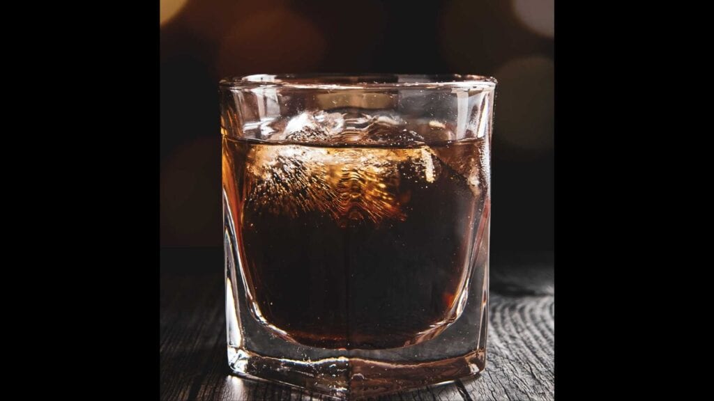 A glass filled with a dark beverage and ice cubes sits on a wooden surface with a blurred background.