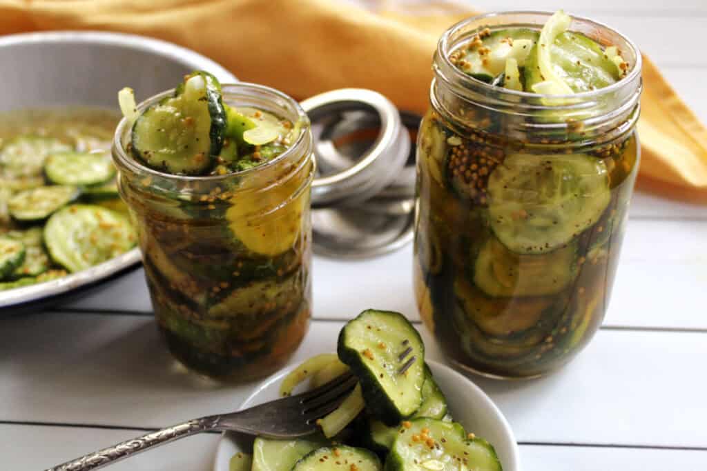 Two jars filled with sliced pickles and onions are on a white surface, with a bowl of additional pickles.