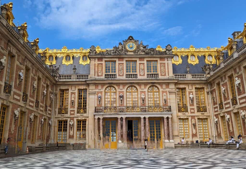 The Palace of Versailles courtyard featuring ornate golden accents, large windows, and detailed sculptures, with people sitting and walking on the checkered floor under a clear blue sky.