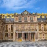 The Palace of Versailles courtyard featuring ornate golden accents, large windows, and detailed sculptures, with people sitting and walking on the checkered floor under a clear blue sky.