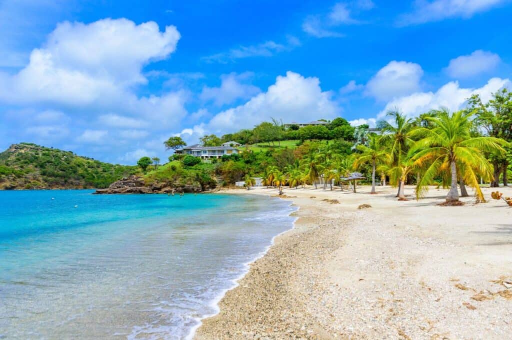 A pristine Caribbean beach with turquoise waters and palm trees, fronted by hillside villas and lush greenery under a partly cloudy sky—truly an enchanting island escape.