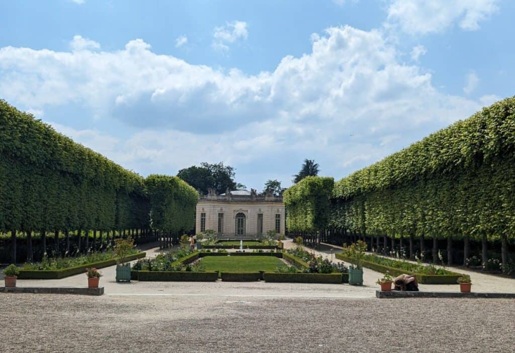 A symmetrical garden path lined with trimmed, green hedges leading to a classical white building under a partly cloudy sky.