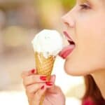 Close-up of a woman licking an ice cream cone. The ice cream is white and the cone is waffle-style. The woman has red nail polish and is viewed from the side against a blurred background.