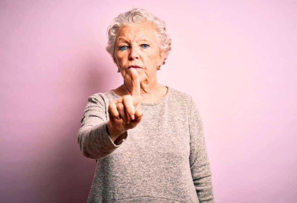 Elderly woman with short white hair stands against a pink background, wearing a gray sweater, raising and pointing her index finger at the camera.