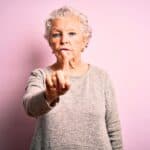 Elderly woman with short white hair stands against a pink background, wearing a gray sweater, raising and pointing her index finger at the camera.