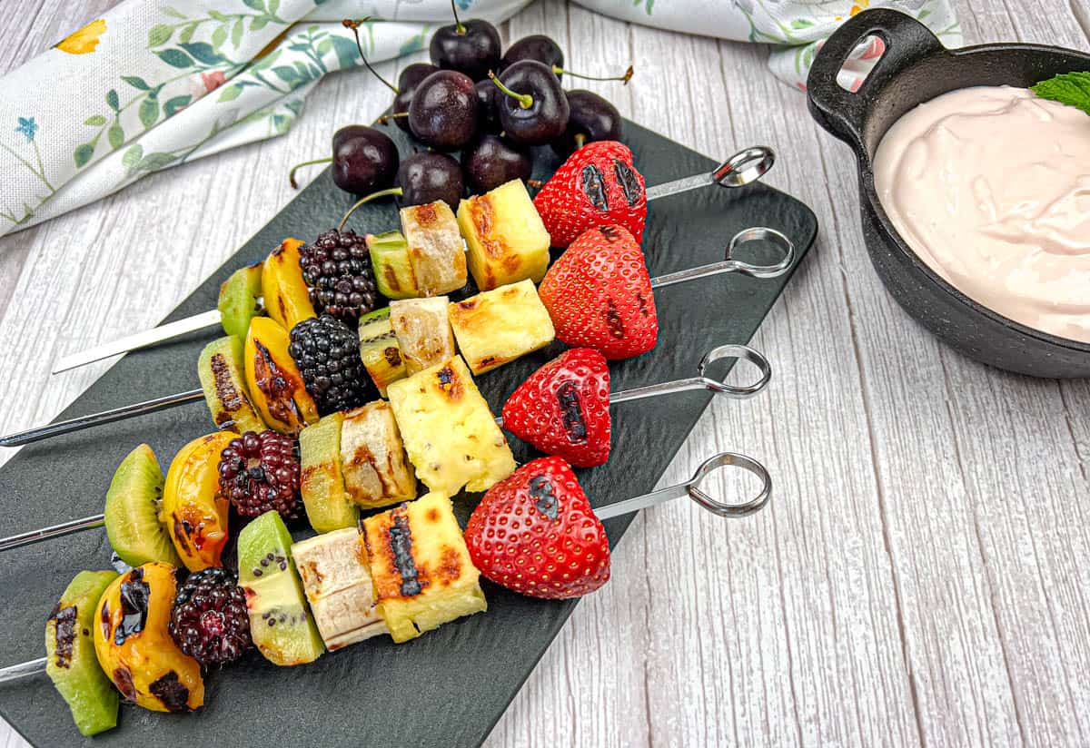 Four pieces of grilled fruit skewers on a slate plate with a bowl of cherry dip.