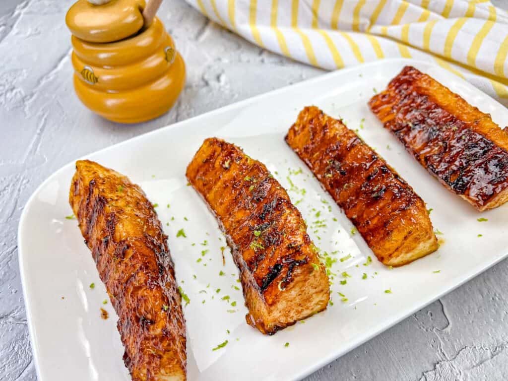 Four grilled salmon fillets glazed with honey and garnished with herbs on a white rectangular plate. A honey pot and a striped cloth are in the background.