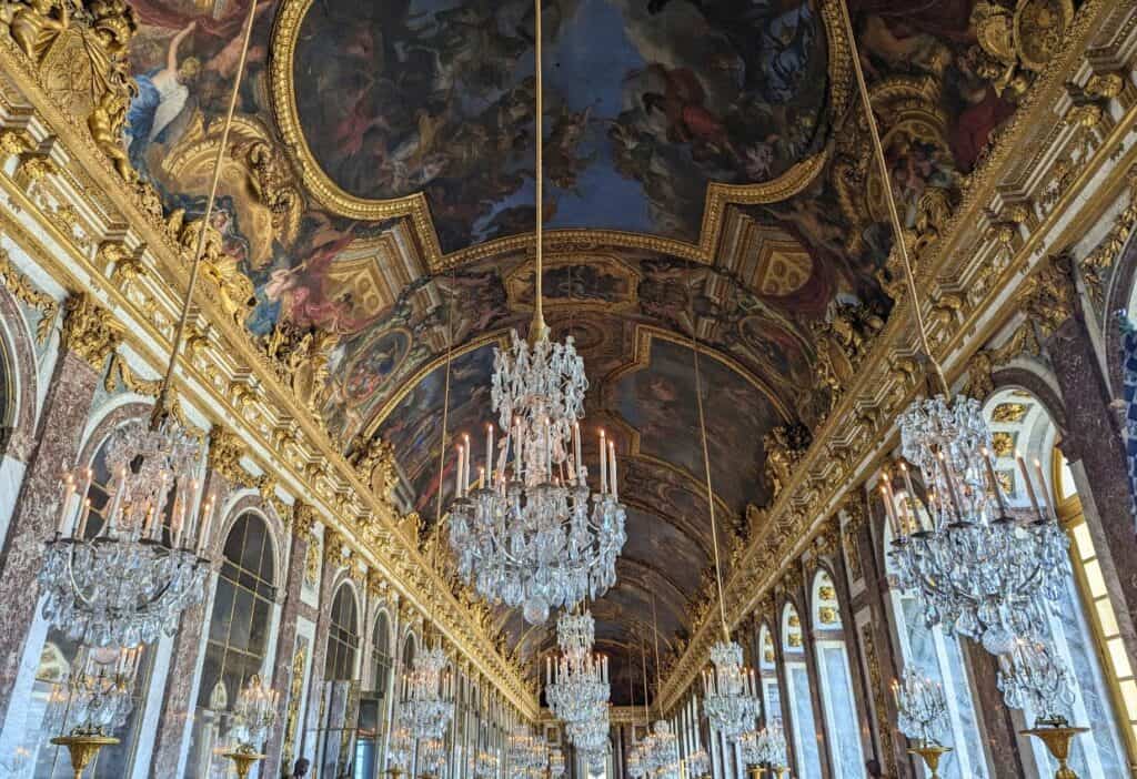 Ornate hall with crystal chandeliers, gilt decorations, and large frescoed ceiling. The room features columns, mirrors, and large windows. People are visible in the background, admiring the architecture.