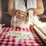 A person in an apron prepares authentic Italian pasta by hand over a red and white checkered tablecloth, with a rolling pin and flour-covered surface nearby, embodying the essence of healthy eating.