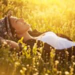 A person lies on their back in a field of grass and wildflowers, wearing a floral hat and white shirt, basking in the warm sunlight, as if absorbing the natural vibrational healing energy from their surroundings.