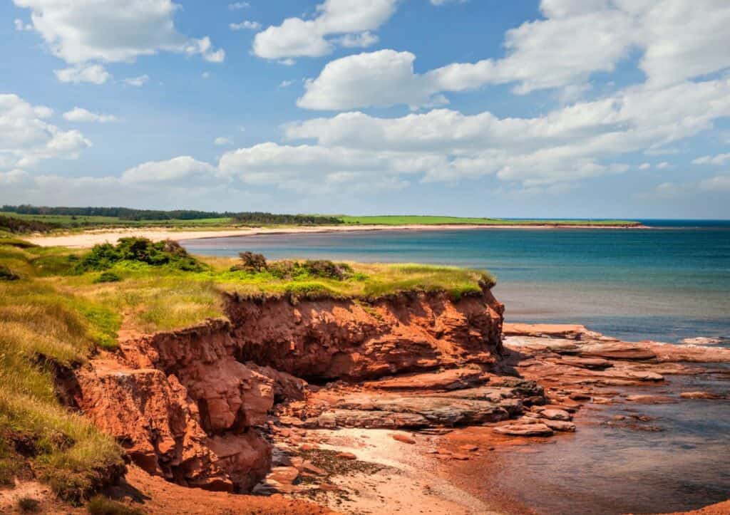 Red sandstone cliffs and green grass overlooking a calm, blue ocean under a partly cloudy sky.