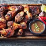 Grilled chicken drumsticks on a wooden tray with a small bowl of dipping sauce, garnished with green onions, two red chili peppers, and a wedge of lemon—all evoking the vibrant flavors of Caribbean best street food—set on a weathered wooden surface.