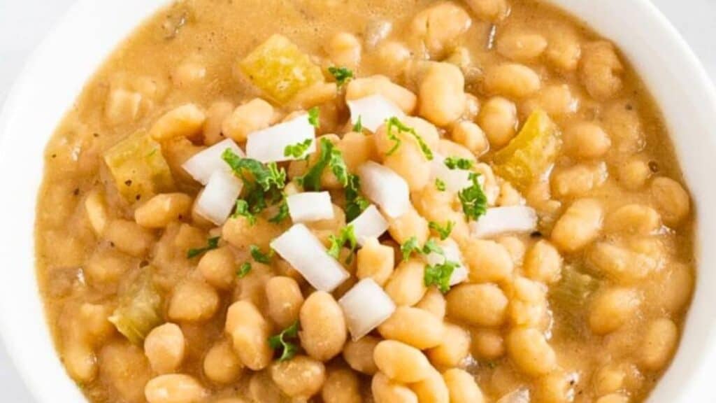 A bowl of bean soup garnished with diced onions and fresh parsley. The soup is thick with visible white beans and pieces of celery.