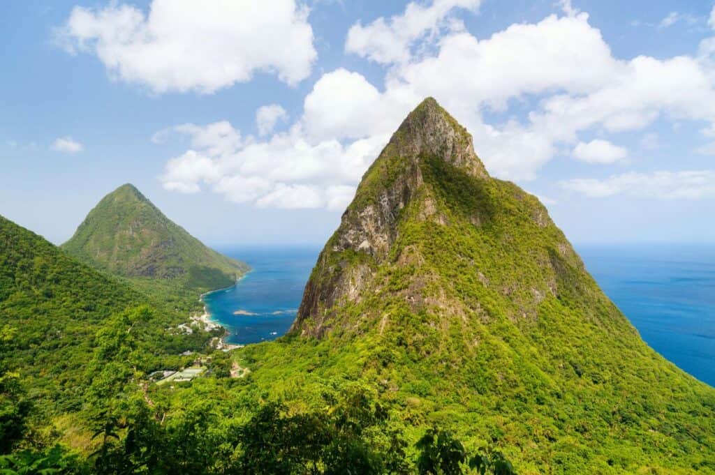 A landscape image featuring two prominent, Piton mountains peaks located near a coastline with clear, blue ocean waters under a partly cloudy sky, reminiscent of scenes from the Caribbean ultimate island guide.