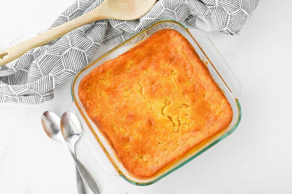 Glass baking dish containing a golden-brown baked cornbread beside two teaspoons and a wooden spoon on a patterned cloth.